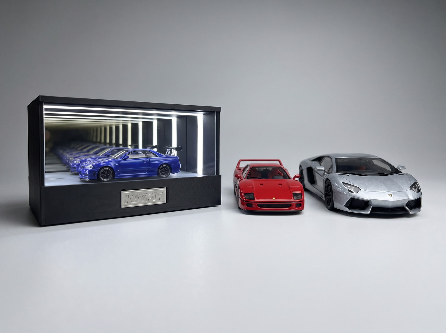 A 1:64 diecast in the lit KP Design Infinity Garage next to a larger 1:43 and an even larger 1:18 model car showing scale differences