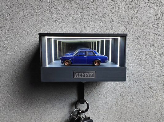 A blue 1:64 Datsun 510 with bronze wheels shown in side profile inside a wall-mounted KP Design Infinity Garage display case