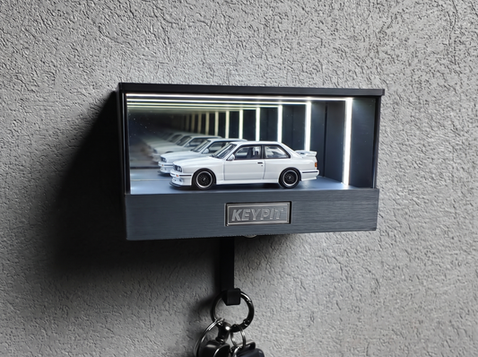A white 1:64 BMW E30 M3 shown in side profile inside a wall-mounted KP Design Infinity Garage display case