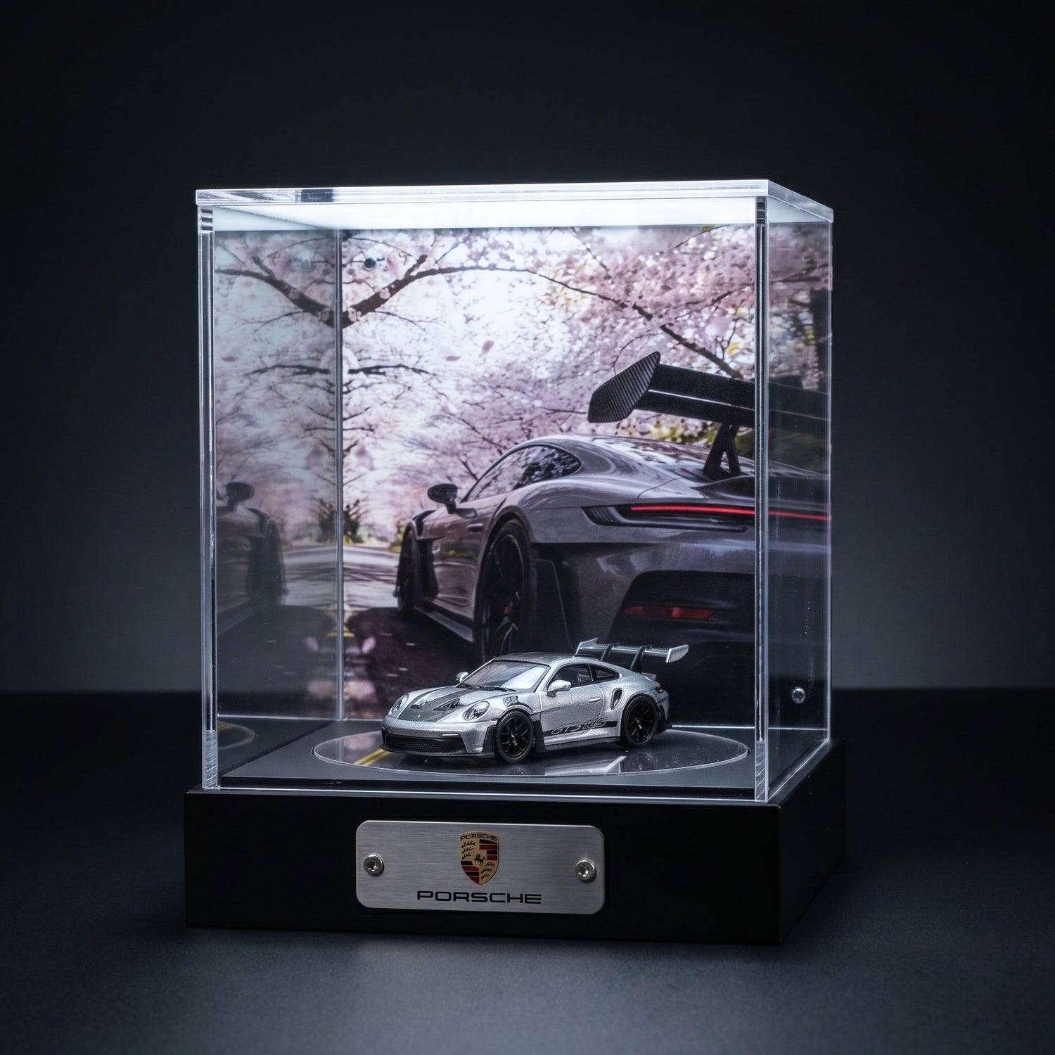 A silver Porsche 911 GT3 RS 1:64 in a lit KP Design Rotating Stage with a cherry blossom backdrop and Porsche nameplate